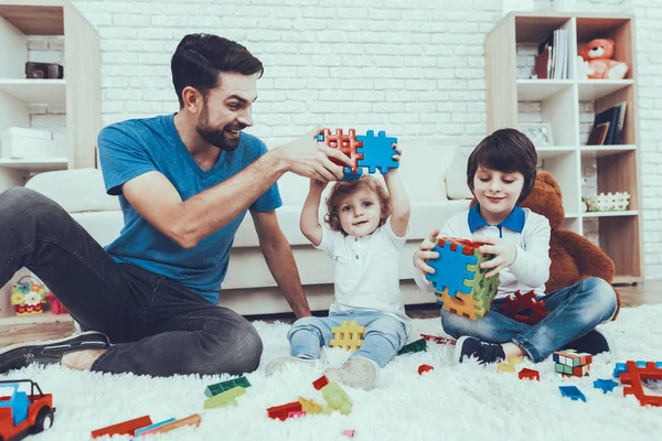 Man Spends Time with His Sons. Father is Engaged in Raising Children. Father and Sons is Playing with Toys. Toys is Lego. Persons is Smiling. Persons is Sitting on Carpet. People Located in Bedroom.