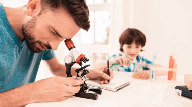 Youn evde baba öğretim oğul gömlekli sakallı. Evde eğitim. Mikroskop kullanarak. Öğrendiğim kimya. Oda beyaz tablosunda. Oturan çocuk. Genç baba. Evde ders. Eğitim kavramı.