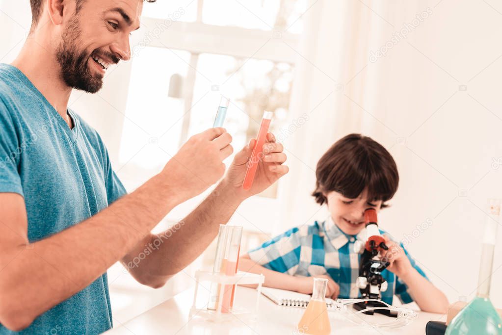 Youn Bearded Father Teaching Son in Shirt at Home. Education at Home. Using Microscope. Studding Chemistry. White Table in Room. Sitting Boy. Young Father. Lesson at Home. Education Concept.