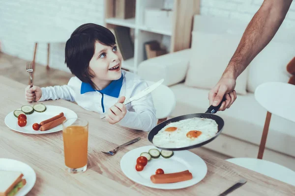 Man Spends Time with His Son. Father of Boy is Engaged in Raising Child. Father is Feeding a Eldest Son a Breakfast. Son is Sitting on Chair at Table and Smiling. People Located on Kitchen.