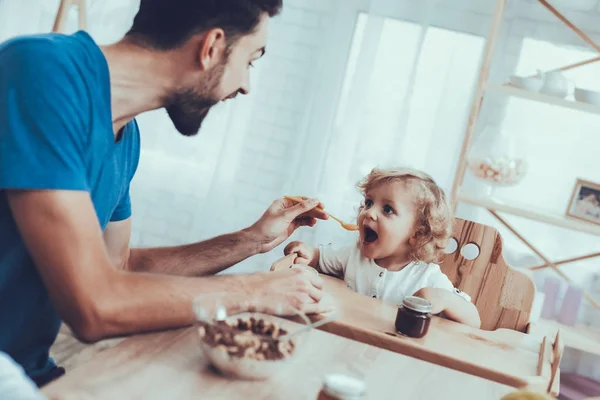 Man Spends Time with His Son. Father of Boy is Engaged in Raising Child. Father is Feeding His Son from Spoon. Man is Smiling. People is Sitting on Chairs at Table. People Located on Kitchen.