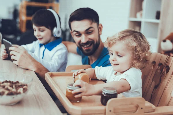 彼の息子との時間を費やしています 父親は子育てに取り組んでいます 長男は タブレット より若い息子にビデオの視聴はベビーフードで遊んでです 男の子は テーブルに座っています キッチンの人 — ストック写真