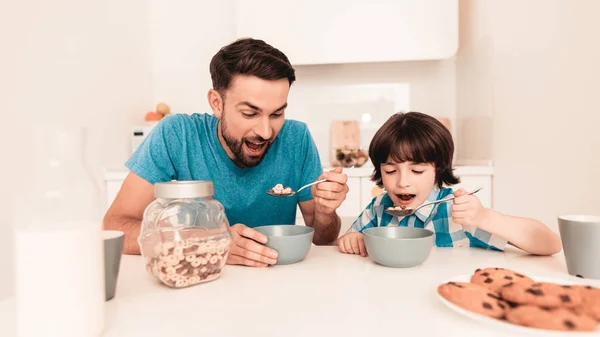 Smiling Son and Father Have Breakfast in Kitchen. Boy in Shirt. Modern Kitchen. Sitting Boy. Boy with Spoon. Breakfast in Morning. White Table in Kitchen. Gray Bowl on Table. Young Father.