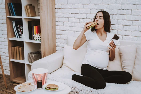 Woman is Eating a Hamburger and Chocolate Bar. Woman is a Young Brunette Pregnant Girl. Girl is Sitting on Couch. Different Food and Cola on Table. Person is Located at Home Interior.