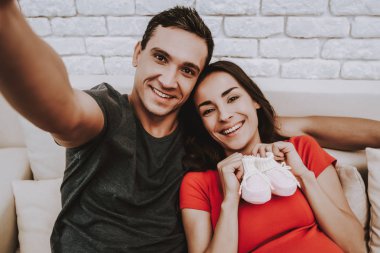 Çift yapıyor Selfie cep telefonu var. Çift hamile kadın ve erkek olduğunu. Kız çocuk ayakkabı tutuyor. Gülen kişidir. Kocam sarılma karısı değil. İnsanların kanepesinde oturuyor. Evde insanlar.