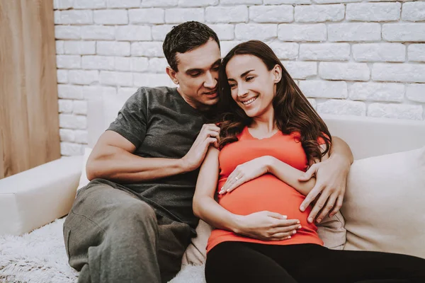 男人正在拥抱他怀孕的妻子 妻子是一个年轻的褐发女郎 丈夫是支持一个女人 一对夫妇是微笑 女孩在摸她的肚子 人们坐在沙发上 在家中的人 — 图库照片