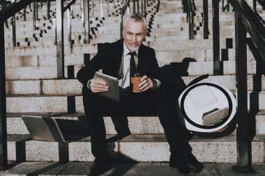 İşadamı bir Tablet Pc. işadamı kullanarak eski gülümseyen adam adam bir fincan kahve Holding. Siyah takım elbise giyen adam. İşadamı merdiven üzerinde oturuyor. Monowheel yakınındaki bir adam güneşli gündüz.
