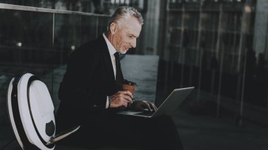 İşadamı bir dizüstü bilgisayar kullanıyor. Eski gülümseyen adam adam giymiş siyah elbiseli işadamıdır. Bir kahve fincanı tutan adam. İş adamı çatıda oturuyor. Monowheel yakınındaki bir adam güneşli gündüz.