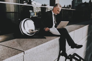 İşadamı bir dizüstü bilgisayar kullanıyor. Eski gülümseyen adam adam giymiş siyah elbiseli işadamıdır. Bir gazete tutan adam. İş adamı çatıda oturuyor. Monowheel yakınındaki bir adam güneşli gündüz.