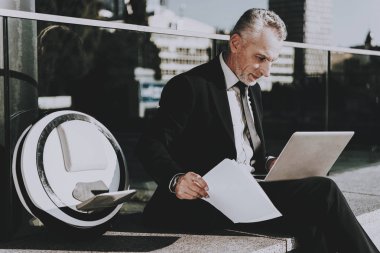 İşadamı bir dizüstü bilgisayar kullanıyor. Eski gülümseyen adam adam giymiş siyah elbiseli işadamıdır. Bir gazete tutan adam. İş adamı çatıda oturuyor. Monowheel yakınındaki bir adam güneşli gündüz.