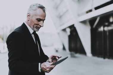 İşadamı bir Tablet Pc. işadamı kullanarak eski gülümseyen adam işadamı Typing tablet olmasıdır. Giyim siyah takım elbiseli biri. Adam ayakta gökdelen yakınındaki yalnızdır. Güneşli gün.