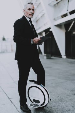 İş adamı olan kullanma bir Tablet Pc. eski gülümseyen adam adam giymiş siyah elbiseli işadamıdır. Gökdelen yakınındaki Monowheel üzerinde duran adam. Adam Uzaklik arıyor. Güneşli gün.
