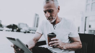 Biri kullanma bir Tablet Pc. adam Tablet ekranda arıyor. Eski gülümseyen adam kişi koltukta oturuyor. Kişi kahve bardağı götürüyor. Güneşli gündüz. İş merkezleri arasında bulunan kişi.