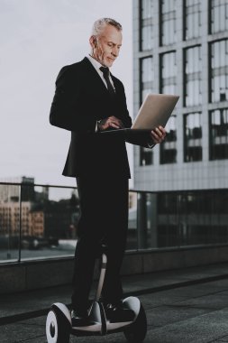 İşadamı Gyroboard biniyor. Kullanma ve bir dizüstü bilgisayarda Typing adamdır. Eski gülümseyen adam adam giymiş siyah elbiseli işadamıdır. İşadamı gökdelenin çatı biniyor. Güneşli gün.
