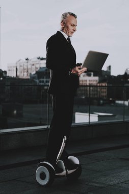 İşadamı Gyroboard biniyor. Kullanma ve bir dizüstü bilgisayarda Typing adamdır. Eski gülümseyen adam adam giymiş siyah elbiseli işadamıdır. İşadamı gökdelenin çatı biniyor. Güneşli gün.