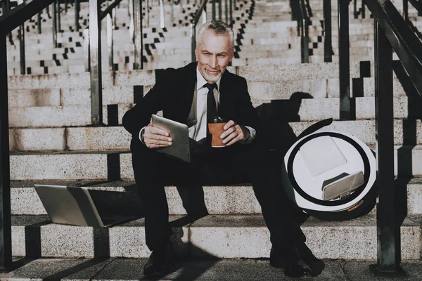 İşadamı bir Tablet Pc. işadamı kullanarak eski gülümseyen adam adam bir fincan kahve Holding. Siyah takım elbise giyen adam. İşadamı merdiven üzerinde oturuyor. Monowheel yakınındaki bir adam güneşli gündüz.
