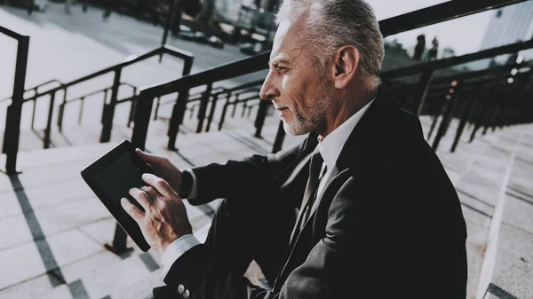 Businessman is Using a Tablet PC. Businessman is Old Smiling Man. Man is Looking on Tablet Screen. Man Wearing in Black Suit. Businessman is Sitting on Staircase of Skyscraper. Sunny Daytime.