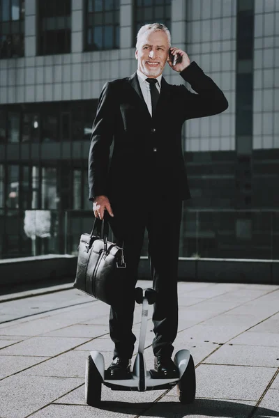İşadamı Gyroboard biniyor. Eski gülümseyen adam adam giymiş siyah elbiseli işadamıdır. Adam bir evrak çantası tutuyor. Cep telefonunda Talking işadamıdır. Gökdelenin çatı. Güneşli gün.