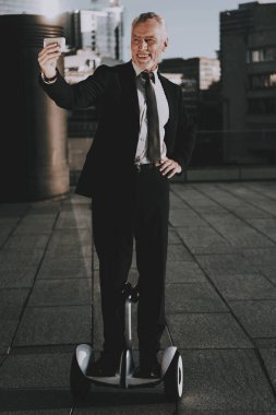 İşadamı Gyroboard biniyor. Adam yapıyor Selfie cep telefonu var. Eski gülümseyen adam adam giymiş siyah elbiseli işadamıdır. İşadamı gökdelenin çatı biniyor. Güneşli gün.