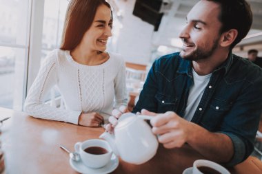 Cafe dinlenme çift. İki güzel genç erkek ve kadın var. Adam bir çay üzerinden su ısıtıcısı için kadın yağıyor. Kişi masada oturuyor. Mutlu ve gülen kişidir. Güneşli gün.