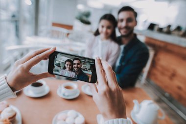 Aile Cafe dinleniyor. Anne, baba ve kızı ailedir. Anne fotoğrafları baba kızı üstünde hareket eden telefon ile. Kişi masada oturuyor. Mutlu ve gülen kişidir. Gündüz.