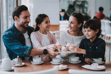 Aile Cafe dinleniyor. Anne, Baba, kızı ve oğlu ailedir. Yemek kek ve bir çay içme kişidir. Kişi masada oturuyor. Mutlu ve gülen kişidir. Güneşli gün.