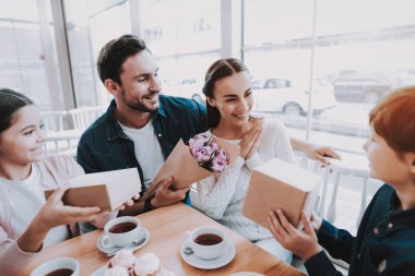 Aile Cafe bir tatil kutluyor. Anne, Baba, kızı ve oğlu ailedir. Baba ve çocuk bir çiçek vererek ve hediyeler anne. Mutlu ve gülen kişidir. Kek ve çay tablo.