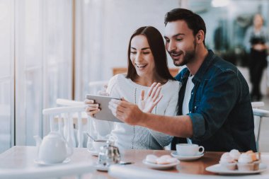 Cafe dinlenme çift. İki güzel genç erkek ve kadın var. Çift Video seslenmek Tablet Pc. var kişi masada oturuyor. Mutlu ve gülen kişidir. Kek ve çay tablo. Gündüz.