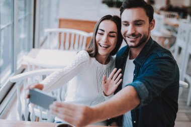 Cafe dinlenme çift. İki güzel genç erkek ve kadın var. Çift yapıyor Selfie cep telefonu var. Kişi masada oturuyor. Dalgalanan bir kadındır. Mutlu ve gülen kişidir. Gündüz.