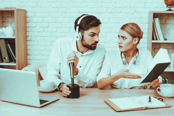 Bloggers Makes a Video. Bloggers is Surprised Man and Woman. Camera Shoots a Video. People Wearing a Headphones. Woman Showing a Notebook to Man. Microphone and Laptop on Table. Studio Interior.