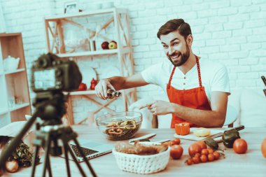 Blogger bir Video yapar. Blogger gülümseyen sakallı adam Video hakkında bir yemek olduğunu. Kamera bir Video vuruyor. Laptop ve farklı yemek masada. Dökülen petrol salata adamdır. Studio iç adam.
