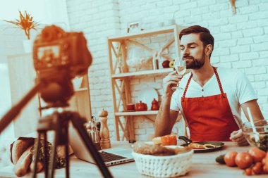 Blogger bir Video yapar. Blogger Genç sakallı adam Video hakkında bir yemek olduğunu. Kamera bir Video vuruyor. Laptop ve farklı yemek masada. Bir beyaz şarap kadehinin koklama erkek. Studio iç adam.