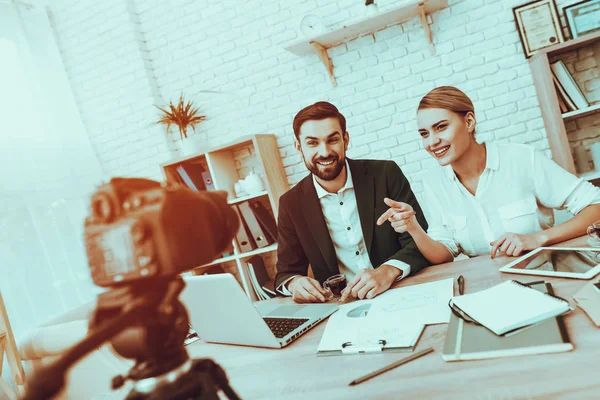 Bloggers Makes a Video. Bloggers is Young Smiling Man and Woman. Video About a Business. Camera Shoots a Video. Gadgets and Supplies on Table. People Looking into a Camera. People in Studio Interior.