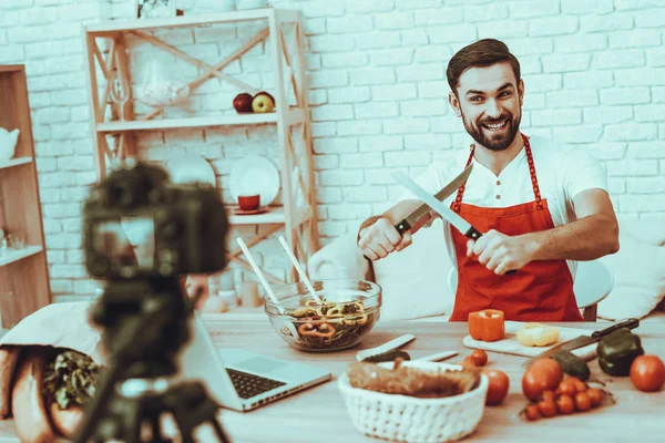 Blogger Makes a Video. Blogger is Smiling Beard Man. Video About a Cooking. Camera Shoots a Video. Laptop and Different Food on Table. Man Showing a Knives. Man in Studio Interior.