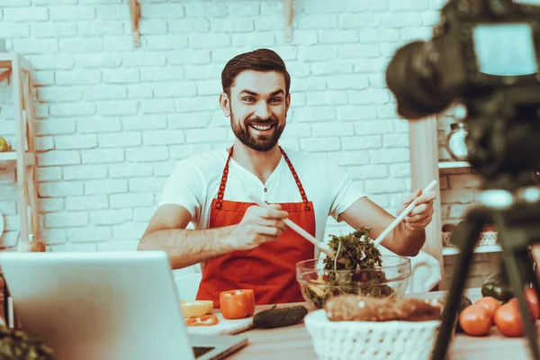 Blogger Makes a Video. Blogger is Smiling Beard Man. Video About a Cooking. Camera Shoots a Video. Laptop and Different Food on Table. Man Mixing a Salad in Bowl. Man in Studio Interior.