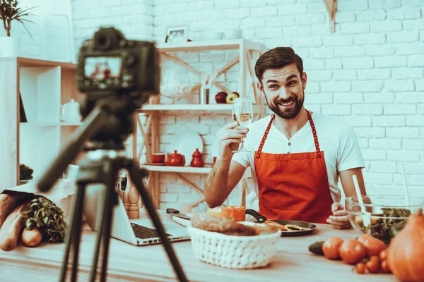 Blogger Makes a Video. Blogger is Smiling Beard Man. Video About a Cooking. Camera Shoots a Video. Laptop and Different Food on Table. Man Showing a White Wine in Wineglass. Man in Studio Interior.