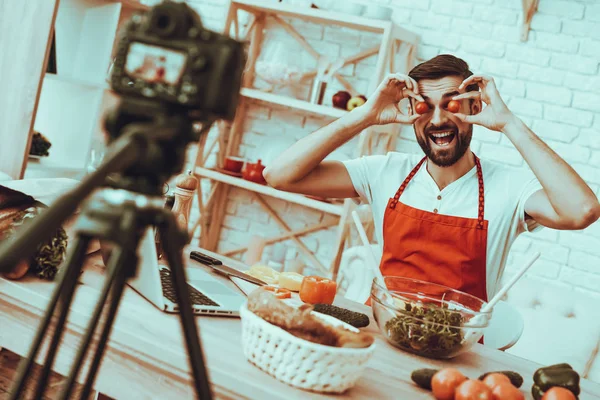 Blogger Makes a Video. Blogger is Smiling Beard Man. Video About a Cooking. Camera Shoots a Video. Laptop and Different Food on Table. Man Showing a Tomatoes. Man in Studio Interior.