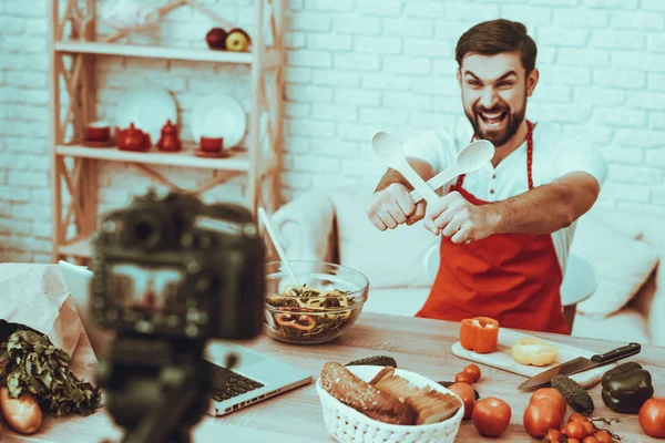 Blogger bir Video yapar. Blogger gülümseyen sakallı adam Video hakkında bir yemek olduğunu. Kamera bir Video vuruyor. Laptop ve farklı yemek masada. Bir tahta kaşıklar gösterilen adam. Studio iç adam.