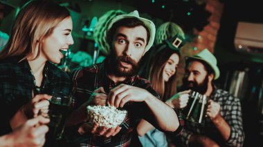 Adam komik şapkalı bir kız ile patlamış mısır yiyor. Bira aperatifler. Alkol kullanımı. Siyah sakallı. Gülümseyen. İyi şenlikli ruh hali. Parlak ışıklar. Club ziyaretçi. Komik şapkalar. Bardakları tokuşturup.