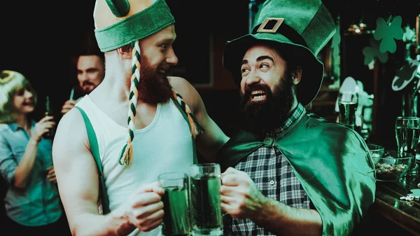 Two Men In Funny Caps. St Patrick\'s Day Concept. Having Fun. Beer Drinking. Bar Counter. Alcohol Handling. Glass Visitors. Bearded Males. Good Festive Mood. Bright Lights. Funny Club Visitors.