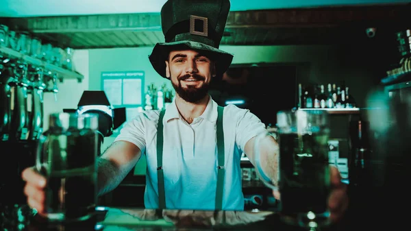 Barman Holds Glasses With Green Beer. St Patrick\'s Day Concept. Clean Bar Counter. Alcohol Handling. Black Beard. Smiling Male. Good Festive Mood. Bright Lights. Club Worker. Funny Hat.