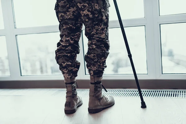 Closeup Soldiers Legs Leaning on Crutch By Window. Disabled Man in High Shoes and Khaki Military Uniform Stands near Large Panoramic Window in Bright Living Room Apartment with Beautiful City View.