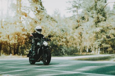 Motosiklet kask içinde bir ormandaki karayolu üzerinde biniyor. Araç hız. Serin binici ile bir deri ceket. Motosiklet kavramı. Klasik tarzı. Sürücü için hazır. Bir ormandaki hız motosiklet. Gizli yüz.