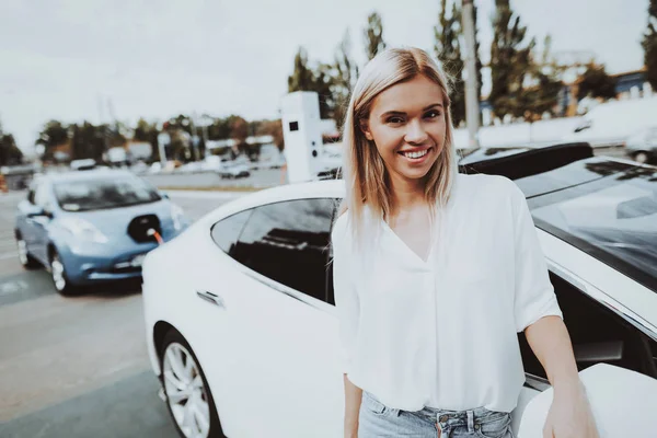 Girl Is Posing On Camera Near Tesla Car In A Charge Station. Automobile Background. Innovation Technology. New Generation Electro Hybrid Vehicle Plugin. Sunny Day. Futuristic Power.