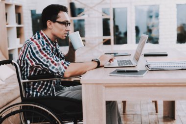 Genç adam evde çalışan tekerlekli sandalye üzerinde devre dışı. Adam tekerlekli sandalye üzerinde. Engelli adam. Kareli gömlek. Ahşap masa. Ahşap raf. Evde serbest meslek. Tekerlekli sandalye üzerinde çalışan. Engelli adam için iş.