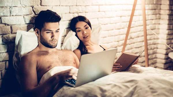 Para przy użyciu laptopa czytanie książki przed snem — Zdjęcie stockowe