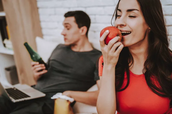 Couple is Sitting on Couch. Woman is Eating an Apple. Woman is a Smiling Young Pregnant Girl. Man is Eating a Chips and Drinking a Beer. Man is Watching Video on Laptop. People Located at Home.
