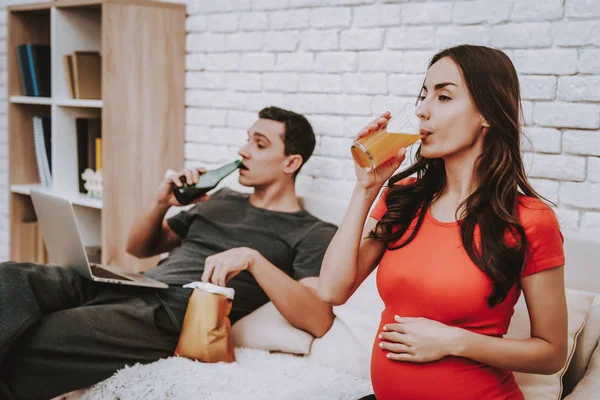 Couple is Sitting on Couch. Woman is Drinking a Juice. Woman is a Young Pregnant Girl. Man is Eating a Chips and Drinking a Beer. Man is Watching Video on Laptop. People Located at Home Interior.