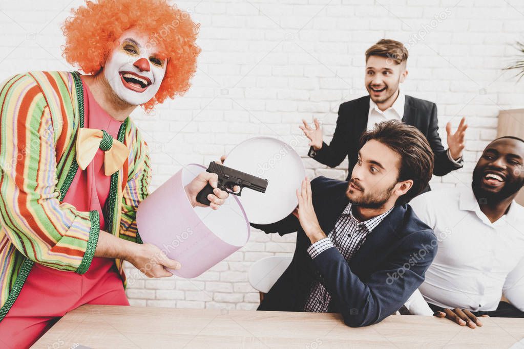 Young Man in Clown Costume on Meeting in Office. April Fools Day. Man with Gift Box. April Jokes. Toy Gun. Workers on Meeting. Holidays and Celebration Concept. Clown with Red Nose.