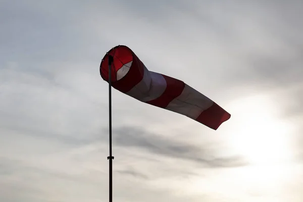 Striped red and white windsock on the pole fluttering in the wind on the cloudy sky and down sun background, closeup